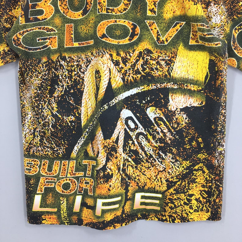 Peut inclure: T-shirt vintage avec un motif graphique avec les mots "BODY GLOVE" en jaune et vert. Le t-shirt porte &eacute;galement les mots "BUILT FOR LIFE" en jaune. Le motif comprend une corde et d'autres &eacute;l&eacute;ments abstraits.