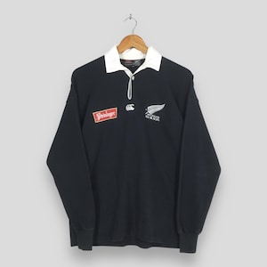 May include: A long-sleeve black rugby shirt featuring a white collar and a button placket. The chest displays a red and white Steinlager logo on the right, a white Canterbury logo in the center, and a white New Zealand All Blacks fern logo on the left.