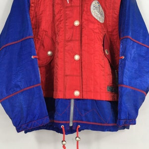 May include: A vintage red and blue jacket with a zip-up front and a drawstring hem. The jacket features a quilted red center panel with large white buttons and a logo patch. The sleeves are blue with red trim.