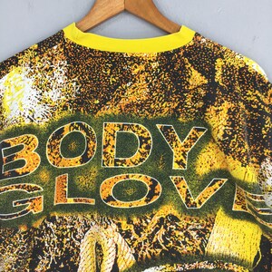 Peut inclure: Un t-shirt vintage &agrave; col rond jaune avec les mots "BODY GLOVE" en grandes lettres soulign&eacute;es. Le t-shirt pr&eacute;sente un motif abstrait vibrant dans les tons de jaune, orange et noir, avec un aspect textur&eacute;.