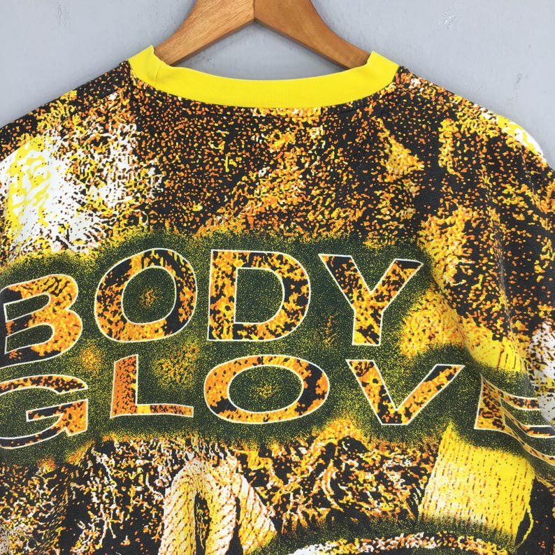 Peut inclure: Un t-shirt vintage &agrave; col rond jaune avec les mots "BODY GLOVE" en grandes lettres soulign&eacute;es. Le t-shirt pr&eacute;sente un motif abstrait vibrant dans les tons de jaune, orange et noir, avec un aspect textur&eacute;.