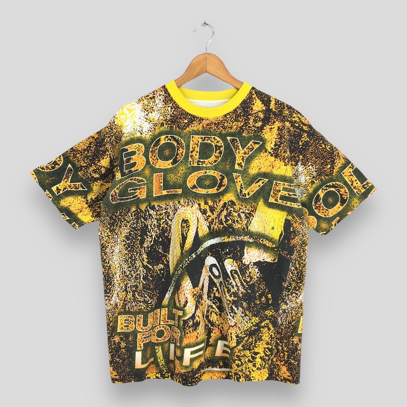 Peut inclure: T-shirt vintage Body Glove avec col jaune et imprim&eacute; int&eacute;gral. Le design pr&eacute;sente les mots "BODY GLOVE" en grandes lettres, ainsi que l'expression "BUILT FOR LIFE". Le t-shirt est suspendu &agrave; un cintre en bois.