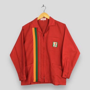 Puede incluir: Una camisa de trabajo roja de manga larga con una franja vertical en verde, naranja y amarillo. La camisa tiene cierre de cremallera, dos bolsillos delanteros y un pequeño logotipo en el pecho. La camisa está colgada en una percha de madera.