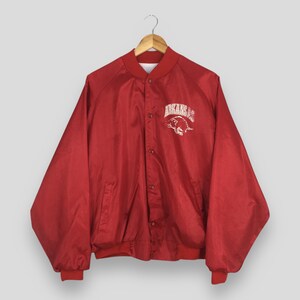 May include: A red satin-finish bomber jacket with a snap-button front closure and long sleeves. The chest features a white embroidered graphic of a hog and the text ARKANSAS. The jacket has a ribbed collar, cuffs, and hem, and is on a hanger.