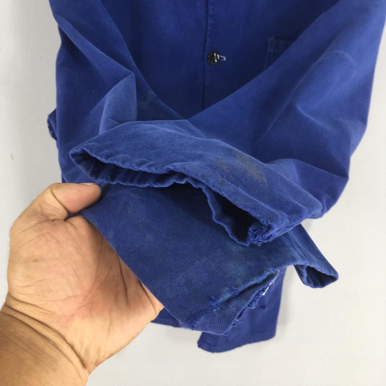 Peut inclure: Gros plan sur une veste de travail bleue. La veste est faite d'un tissu &eacute;pais et durable et comporte une fermeture &agrave; bouton. Les manches sont retrouss&eacute;es et le tissu montre des signes d'usure.
