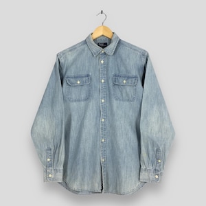 May include: Pale blue denim shirt with long sleeves, two chest pockets, and a button-down front. The shirt is hanging on a wooden hanger. The collar and cuffs have white buttons.