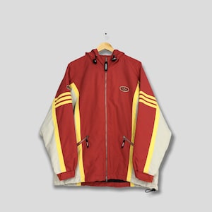 May include: A red, yellow, and cream-colored hooded jacket with a full-length zipper. The jacket features yellow stripes on the sleeves and a small logo on the chest. The jacket is hanging on a wooden hanger.