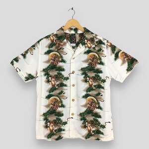 May include: A short-sleeved button-down shirt in white featuring a repeating pattern of green pine branches and small winged figures. The shirt has a classic collar, a front pocket, and light brown buttons. A small black label is visible at the neck.