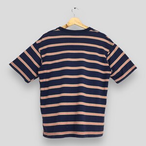 Puede incluir: Camiseta azul marino de manga corta con rayas horizontales rojas y crema. La camiseta est&aacute; colgada de una percha de madera sobre un fondo gris claro. Tiene cuello redondo y un corte holgado.
