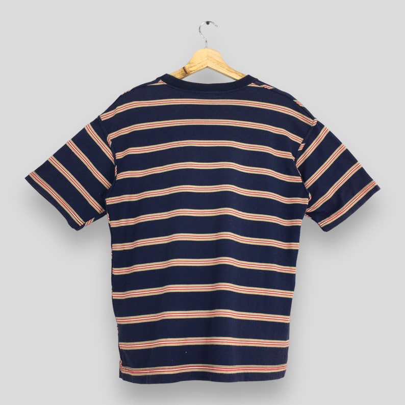 Puede incluir: Camiseta azul marino de manga corta con rayas horizontales rojas y crema. La camiseta est&aacute; colgada de una percha de madera sobre un fondo gris claro. Tiene cuello redondo y un corte holgado.