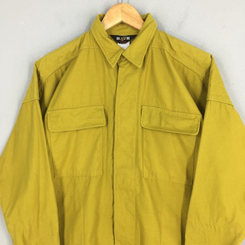 Pode incluir: Camisa mostarda com bot&otilde;es e dois bolsos no peito. A camisa de manga comprida est&aacute; pendurada num cabide de madeira. A camisa tem gola e uma pequena etiqueta perto da gola.