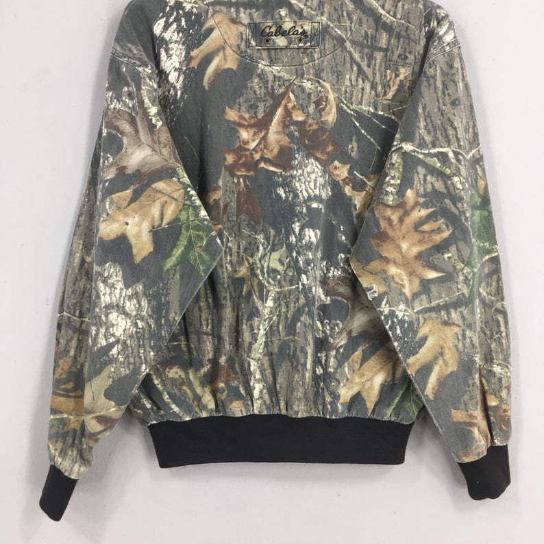 Op de afbeelding: Een sweatshirt met lange mouwen, camouflage-print en een zwarte getailleerde boord. Het sweatshirt heeft het Cabela's logo op de voorkant.