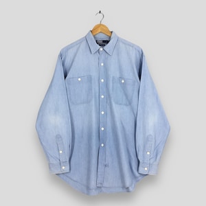 May include: A pale blue long-sleeved shirt with two chest pockets and buttoned cuffs. The shirt is suspended from a wooden hanger. The shirt has a button-down collar and a small logo on the collar.