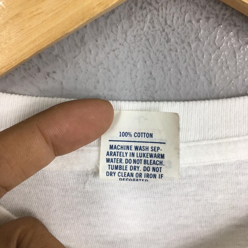 May include: Close-up of a white garment tag with the text "100% COTTON". The tag also includes washing instructions: machine wash separately in lukewarm water, do not bleach, tumble dry, and do not dry clean or iron if decorated.