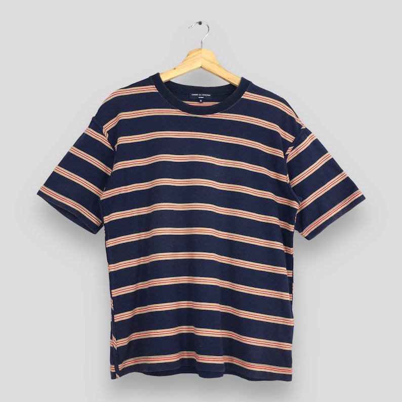 Puede incluir: Camiseta azul marino de manga corta con rayas horizontales en beige y rojo. La camiseta est&aacute; colgada de una percha de madera. El nombre de la marca es visible en la parte superior.