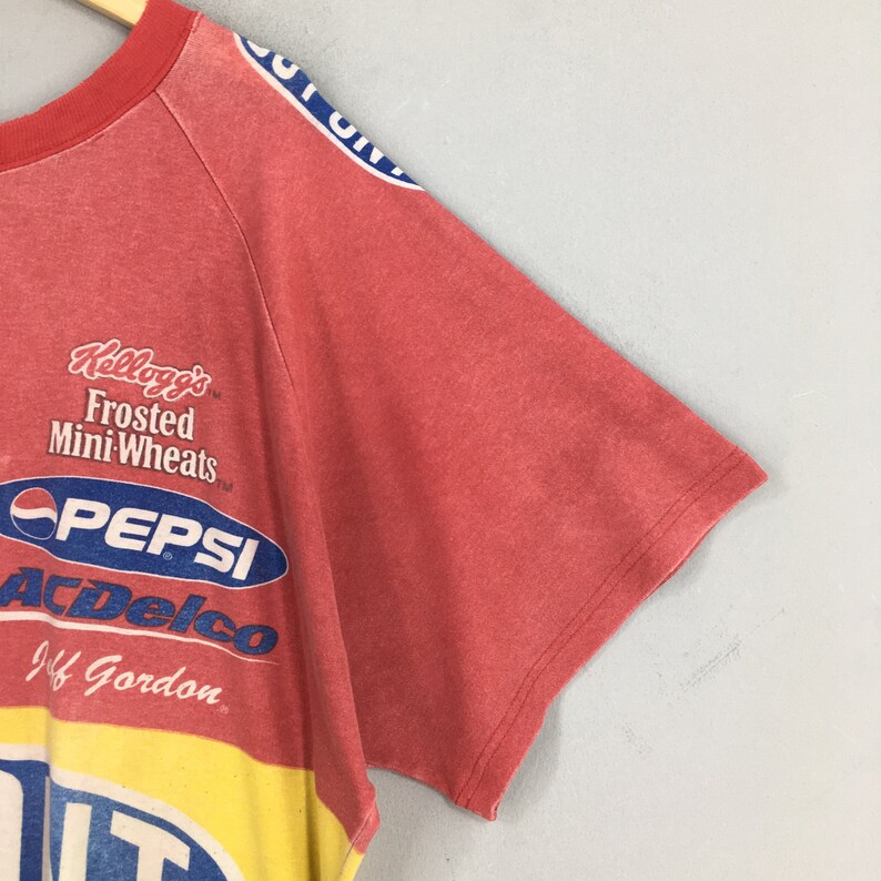 Op de afbeelding: Rood, gele en blauwe vintage t-shirt met sponsorlogo's waaronder Pepsi, Kellogg's Frosted Mini Wheats en ACDelco. De shirt heeft ook de naam Jeff Gordon. De shirt heeft een verweerde uitstraling.