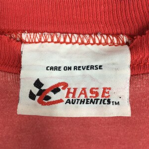 Op de afbeelding: Close-up van een rood kledinglabel met de tekst "CARE ON REVERSE". Het label heeft het "CHASE AUTHENTICS" logo in rood en zwart. Het kledingstuk heeft een effen rode kleur.