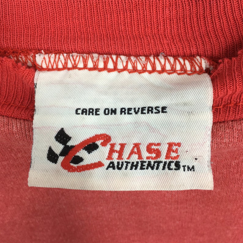 Op de afbeelding: Close-up van een rood kledinglabel met de tekst "CARE ON REVERSE". Het label heeft het "CHASE AUTHENTICS" logo in rood en zwart. Het kledingstuk heeft een effen rode kleur.