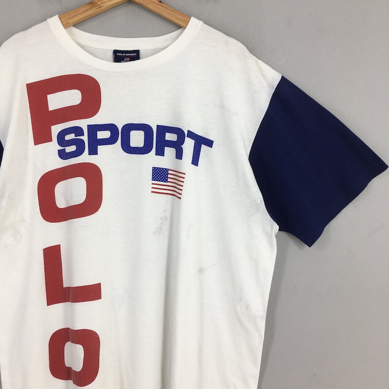 May include: White short-sleeved t-shirt with a red, blue, and white "POLO SPORT" graphic and an American flag. The right sleeve is blue.