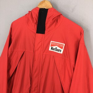 May include: A vibrant red jacket with a hood and a black collar. The jacket displays the "Adventure Team" and "Marlboro" logo on the chest. The jacket has a zip closure and two front pockets.