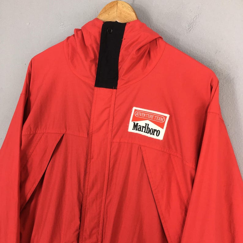 May include: A vibrant red jacket with a hood and a black collar. The jacket displays the "Adventure Team" and "Marlboro" logo on the chest. The jacket has a zip closure and two front pockets.
