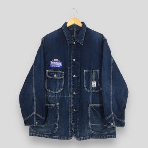 Pode incluir: Um casaco de trabalho em ganga azul escuro com gola pontiaguda, mangas compridas e fecho frontal de botões. Inclui quatro bolsos frontais e um emblema no peito com o texto Rothmans Rally of New Zealand Official e uma pequena etiqueta.