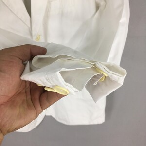 May include: A crisp white long-sleeved shirt featuring a classic collar and a buttoned cuff detail. The garment is constructed with light yellow buttons along the sleeve placket and cuff, providing a subtle contrast against the plain white fabric.