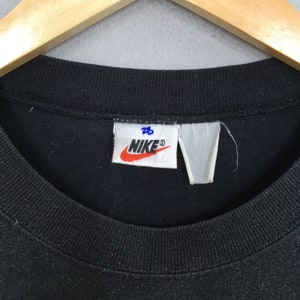 Może przedstawiać: Czarny T-shirt Nike z białą metką, na kt&oacute;rej napisano "Nike" niebieskimi literami i "28" niebieskimi literami.