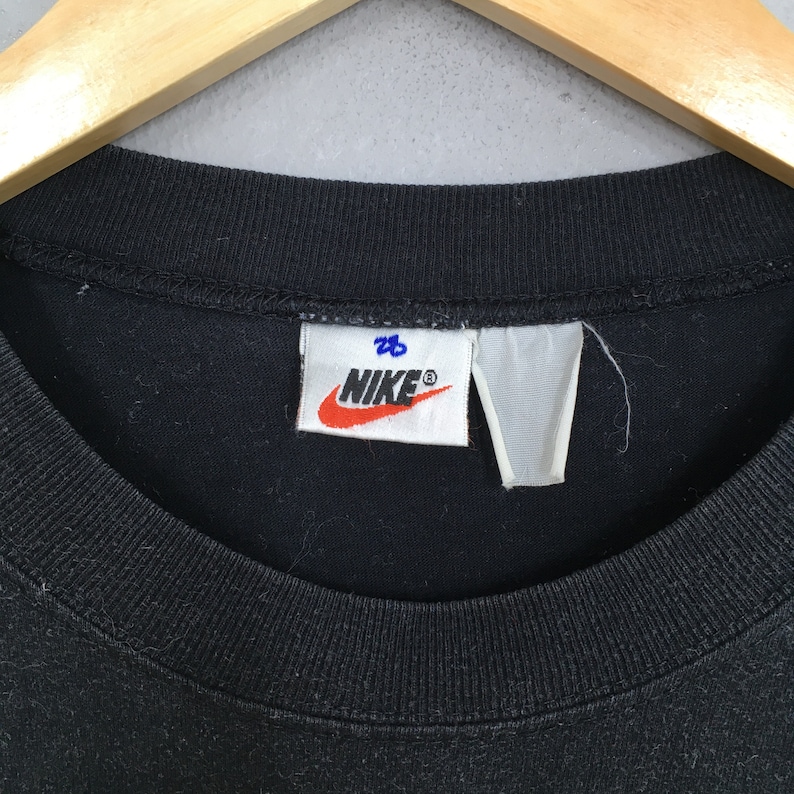 Może przedstawiać: Czarny T-shirt Nike z białą metką, na kt&oacute;rej napisano "Nike" niebieskimi literami i "28" niebieskimi literami.