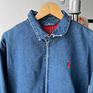 May include: A blue denim jacket with a classic collar and a full-length front zipper. The interior is lined with a red and black plaid fabric. A small red embroidered logo of a person on a horse is visible on the left chest area of the jacket.