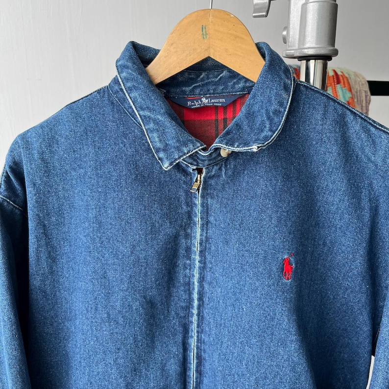 May include: A blue denim jacket with a classic collar and a full-length front zipper. The interior is lined with a red and black plaid fabric. A small red embroidered logo of a person on a horse is visible on the left chest area of the jacket.
