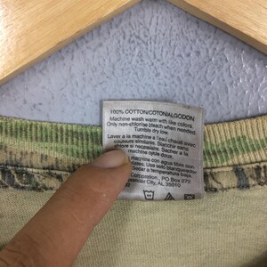 May include: Close-up of a tag with care instructions for a garment. The tag is white with black text, including "100% COTTON" and washing instructions. The tag is attached to a camouflage-patterned fabric.
