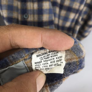 K&ouml;nnte beinhalten: Nahaufnahme eines blau, beige und wei&szlig; karierten Button-Down-Hemds. Das Pflegeetikett ist sichtbar und enth&auml;lt Anweisungen zum Maschinen- oder Handwaschen. Das Etikett empfiehlt auch das Waschen in lauwarmem Wasser.
