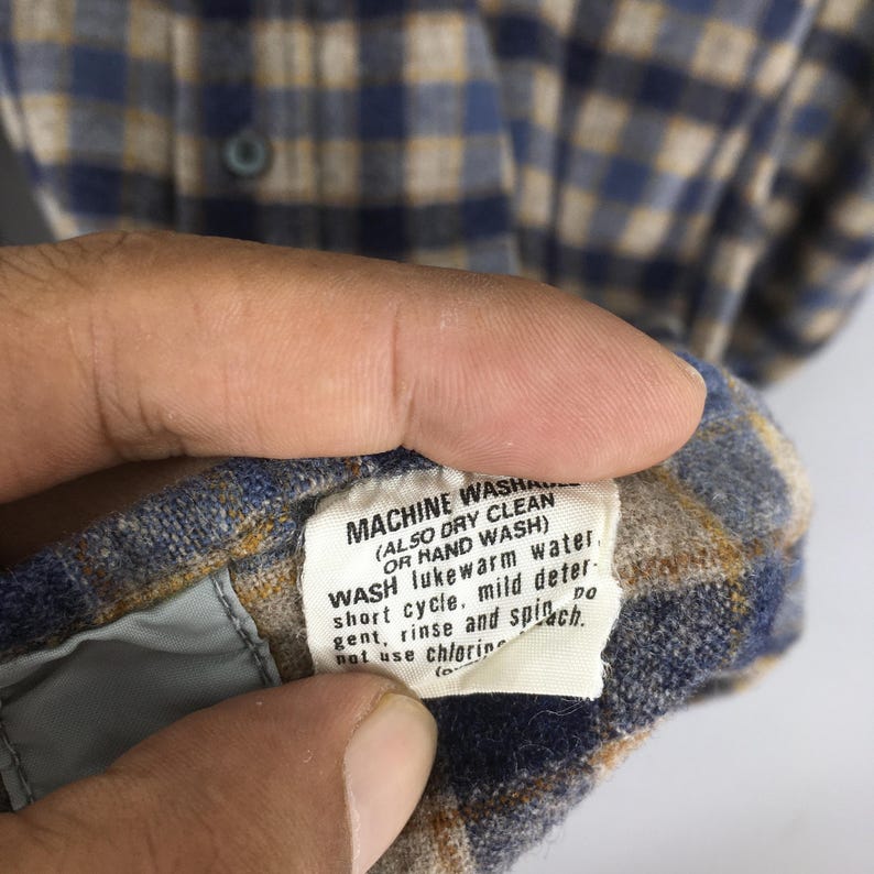 K&ouml;nnte beinhalten: Nahaufnahme eines blau, beige und wei&szlig; karierten Button-Down-Hemds. Das Pflegeetikett ist sichtbar und enth&auml;lt Anweisungen zum Maschinen- oder Handwaschen. Das Etikett empfiehlt auch das Waschen in lauwarmem Wasser.