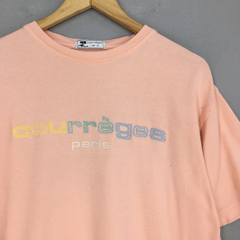 K&ouml;nnte beinhalten: Ein pfirsichfarbenes T-Shirt mit dem gestickten Text "courr&egrave;ges paris" in einem mehrfarbigen Design. Das Shirt h&auml;ngt an einem Holzb&uuml;gel. Das Markenlogo befindet sich oben.