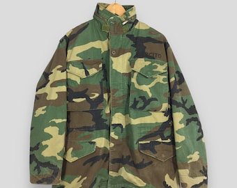 May include: Camouflage pattern jacket with multiple pockets and a high collar. The jacket is in shades of green, brown, and tan. The word "ERCITO" is printed on the right side of the chest. The jacket is hanging on a wooden hanger.