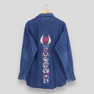 May include: A blue denim long-sleeve shirt with a colorful totem pole design on the back. The design features a pink bird, blue and white faces, and the text "Belle Pelle".