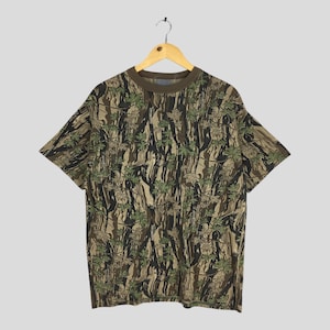 May include: A short-sleeved t-shirt with a camouflage pattern of green leaves and brown and black tree trunks. The shirt has a brown collar and is hanging on a wooden hanger.