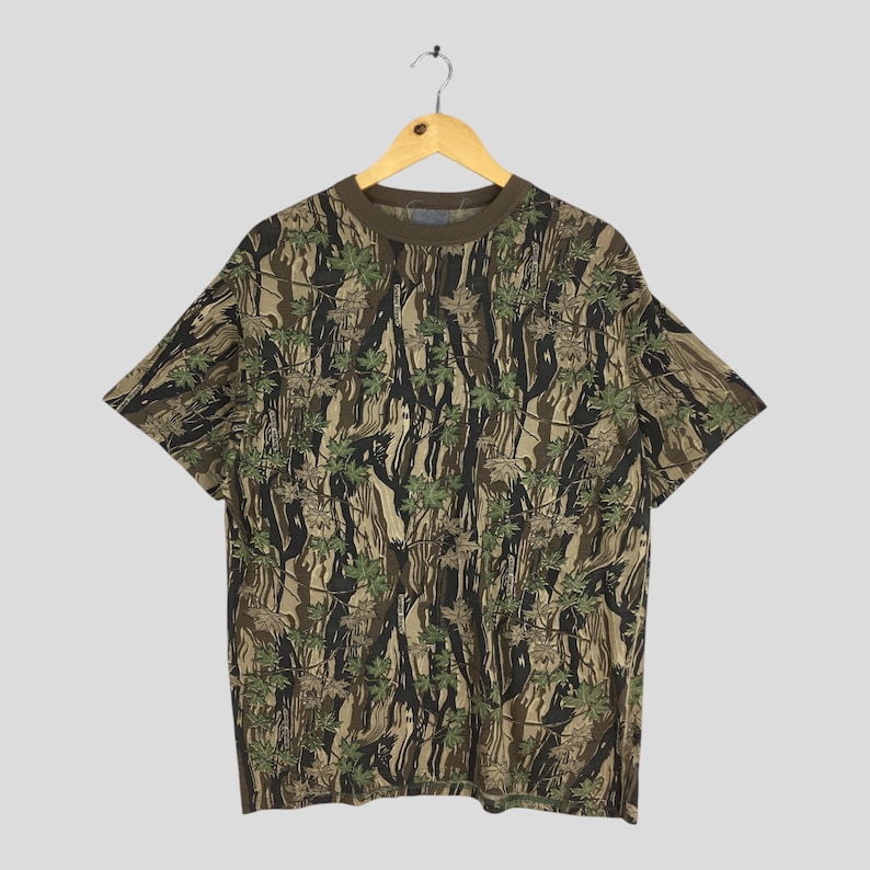 May include: A short-sleeved t-shirt with a camouflage pattern of green leaves and brown and black tree trunks. The shirt has a brown collar and is hanging on a wooden hanger.