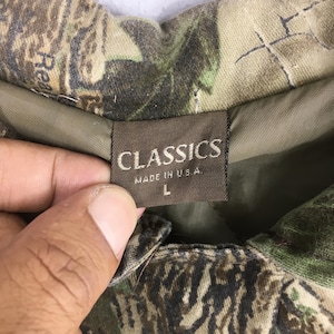 May include: Close-up of a camouflage jacket label. The label reads "CLASSICS MADE IN U.S.A." with a size "L". The jacket has a green and brown camouflage pattern.