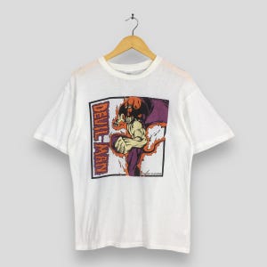 May include: White t-shirt featuring a graphic of a muscular figure with a devilish appearance, flames, and the text "DEVILMAN" in a rectangular frame. The design incorporates shades of red, purple, and yellow.