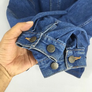 May include: Close-up of a blue denim jacket sleeve. The jacket features metal buttons with a circular design. The denim has visible stitching and a classic, worn appearance. The sleeve is folded, showcasing the cuff and button details.