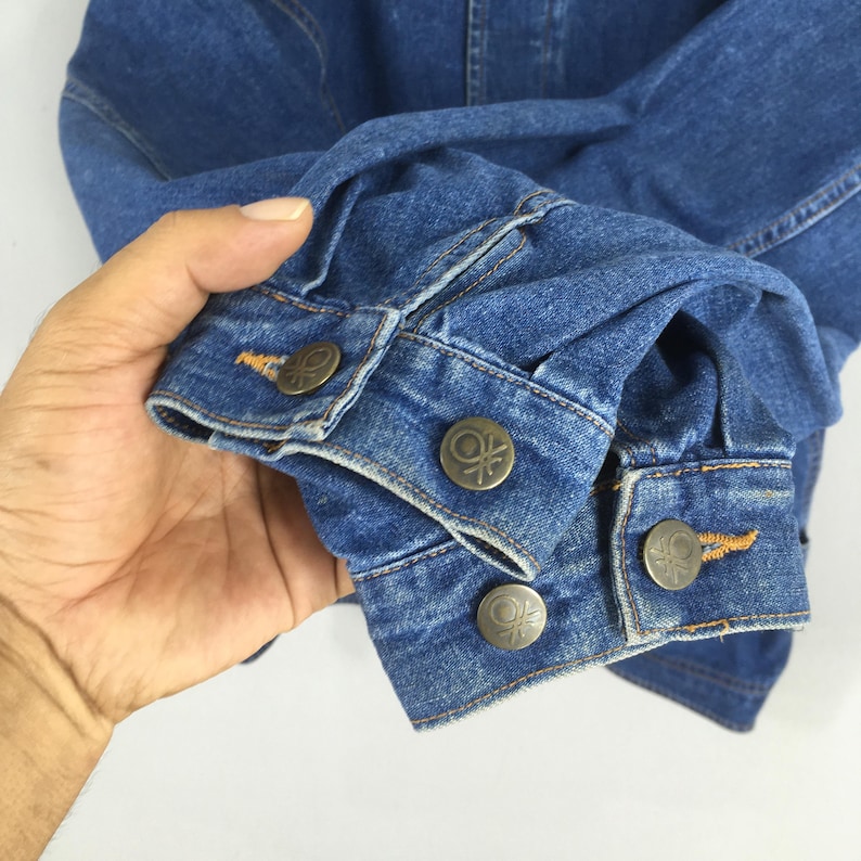May include: Close-up of a blue denim jacket sleeve. The jacket features metal buttons with a circular design. The denim has visible stitching and a classic, worn appearance. The sleeve is folded, showcasing the cuff and button details.