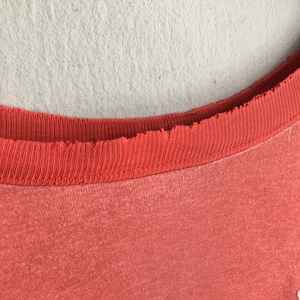 Op de afbeelding: Close-up van een koraalkleurige t-shirt halslijn. De halslijn is een donkerdere tint koraal en heeft een gerafelde, gerafelde rand. De stof van de shirt lijkt zacht en licht vervaagd, met een subtiele textuur.