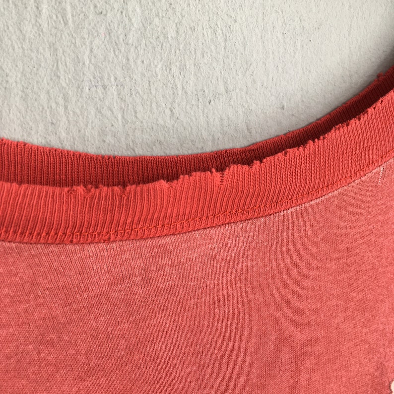 Op de afbeelding: Close-up van een koraalkleurige t-shirt halslijn. De halslijn is een donkerdere tint koraal en heeft een gerafelde, gerafelde rand. De stof van de shirt lijkt zacht en licht vervaagd, met een subtiele textuur.