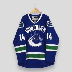 May include: A blue Vancouver Canucks hockey jersey with white and green accents. The jersey features the word "VANCOUVER" in white across the chest, along with the number "14" on the sleeves. The team logo is also visible.