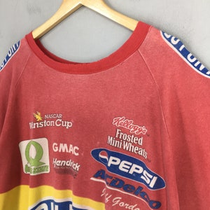 Op de afbeelding: Rode vintage sweatshirt met meerdere logo's, waaronder "NASCAR Winston Cup", "Kellogg's Frosted Mini-Wheats" en "Pepsi". De trui heeft een rode kraag en blauw-witte strepen op de mouwen. De onderkant van de trui is geel.