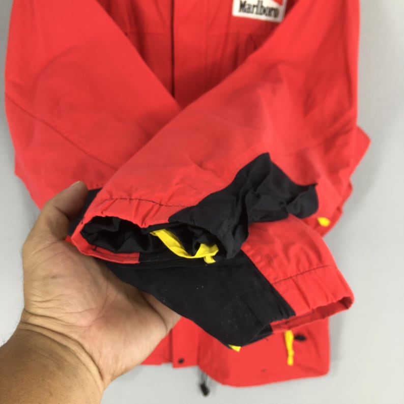May include: Red jacket with black and yellow detailing. The jacket features a Marlboro logo on the upper right. The sleeve cuffs are black with yellow trim. The jacket is made of a lightweight material.