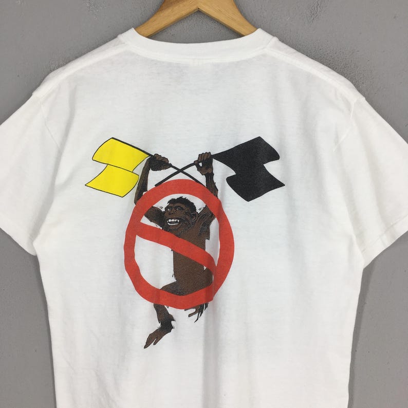 May include: White t-shirt featuring a graphic of a monkey inside a red prohibition sign, holding yellow and black flags. The design is printed on the back of the shirt. The shirt is hanging on a wooden hanger.