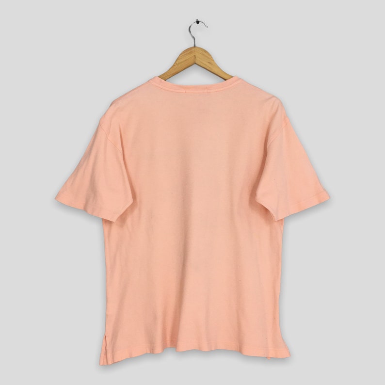 K&ouml;nnte beinhalten: Ein pfirsichfarbenes T-Shirt h&auml;ngt an einem Holzb&uuml;gel vor einem hellgrauen Hintergrund. Das kurz&auml;rmelige Shirt hat einen Rundhalsausschnitt und eine lockere Passform. Der Stoff scheint aus weicher Baumwolle oder einer Baumwollmischung zu sein.